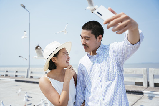 快乐的亚泰夫妇在海边大桥用智能手机拍照。海鸥在背景上飞翔。