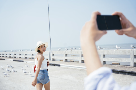 男人拿着智能手机的手拍着女人在海边旅行戴着帽子的女人。