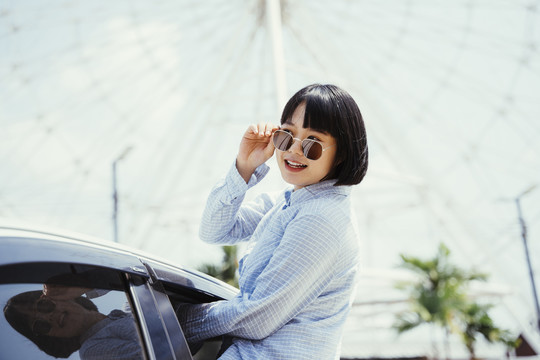 年轻的亚洲女孩鲍勃发型戴着墨镜坐在车窗座位上挥手在车外。