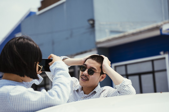一对戴着墨镜的浪漫亚泰情侣在车外对视。女人用智能手机给光头男拍照。