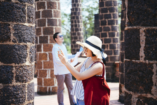 美丽的亚泰游客戴着帽子，戴着墨镜，手持智能手机漫步古城观光。走在燃烧的柱子之间。