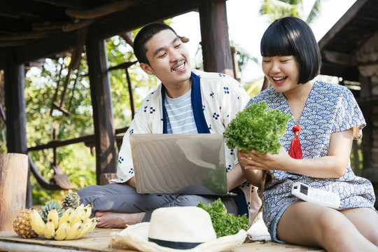 生态旅游——一对亚洲年轻商人在乡村用笔记本电脑卖当地农场的蔬菜。现代农民利用技术销售产品。
