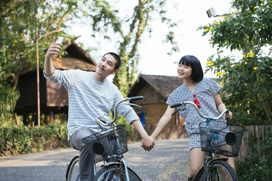 生态旅游——亚洲情侣手牵手骑自行车用智能手机自拍。