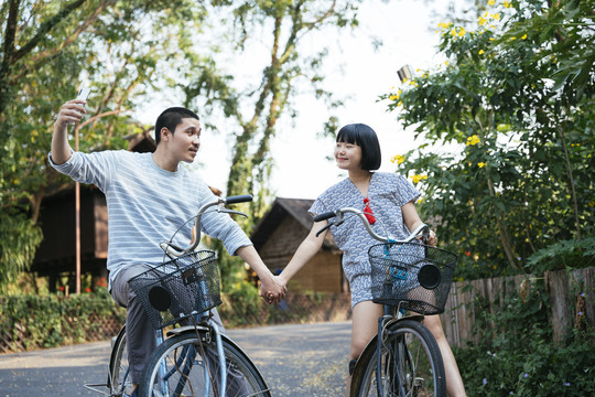 生态旅游——亚洲情侣手牵手骑自行车用智能手机自拍。