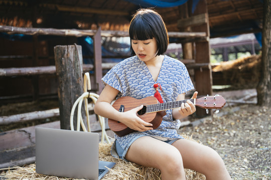 生态旅游-亚泰旅行者鲍勃发型女子在动物农场玩四弦琴吉他，看着笔记本电脑屏幕。乡下女孩。