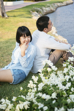 一对浪漫的亚泰夫妇背靠背地坐在河边公园的户外绿地上。前景是白色的花。