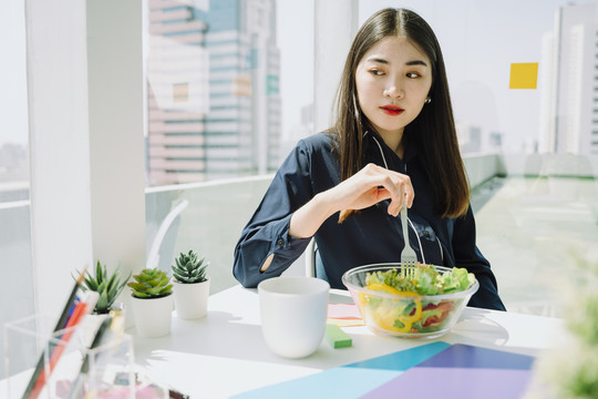 美丽的亚洲年轻泰国女子留着长长的黑发在办公室工作，吃着沙拉碗。健康食品概念。女商人和健康沙拉碗。