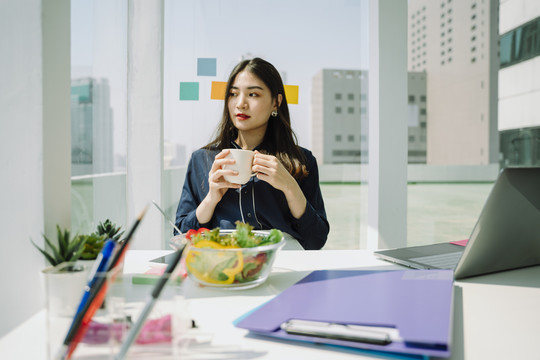 美丽的亚洲年轻泰国女子，一头乌黑的长发，在办公桌边吃午餐。