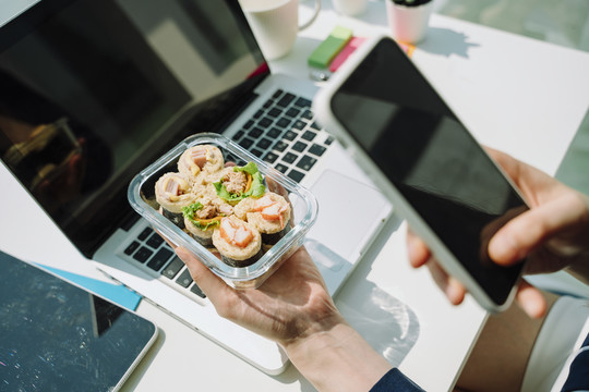 女商人在办公桌上使用笔记本电脑时吃面包春卷的手。用智能手机拍照。