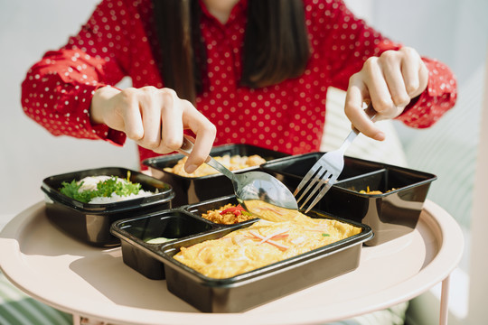 美丽的亚泰长黑发红衫女子拿着用叉子和勺子吃食物的饭盒套装。