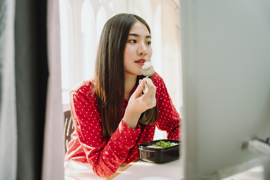 美丽的亚泰黑发红衫女子在家里电脑前吃健康食品。