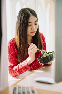 美丽的亚泰黑发红衫女子在家里电脑前吃健康食品。