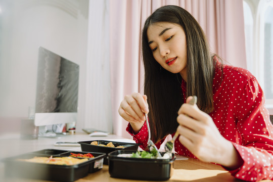 美丽的亚泰黑发红衫女子在电脑前享受健康饮食。在家工作的概念。