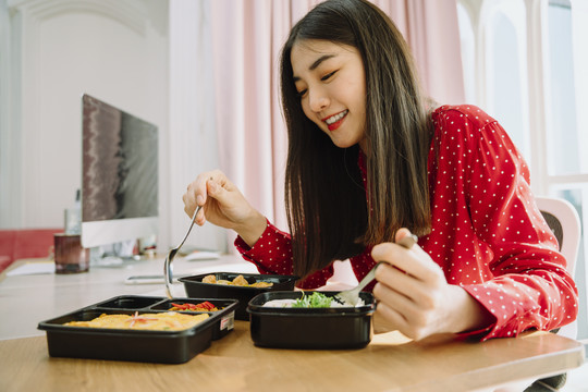 美丽的亚泰黑发红衫女子在电脑前享受健康饮食。在家工作的概念。