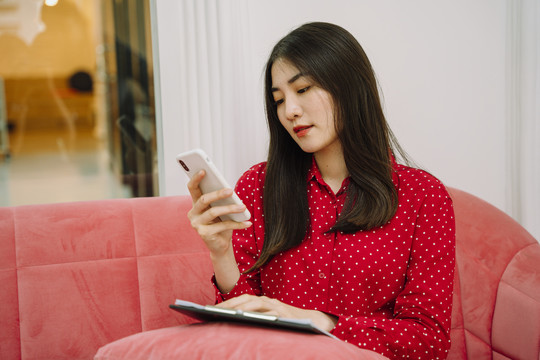 美丽的亚泰黑发红衫女子坐在粉色沙发沙发上，用智能手机与顾客联系，核对纸质工作清单。在家工作。在家工作