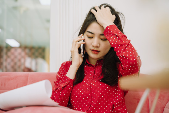 美丽的亚泰黑发红衫女子坐在粉色沙发沙发上，用智能手机与顾客联系，核对纸质工作清单。在家工作。在家工作