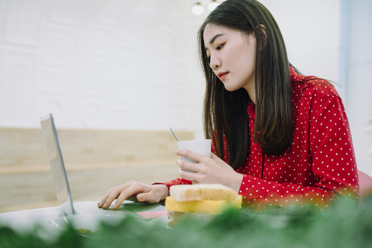 美丽的亚泰黑发长红衫女子端着一杯咖啡，在家里用笔记本电脑工作。在家工作。