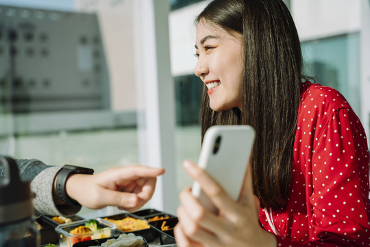 兴高采烈的亚洲红衫女子手持智能手机向朋友展示挑选食物的申请。送货食品申请。