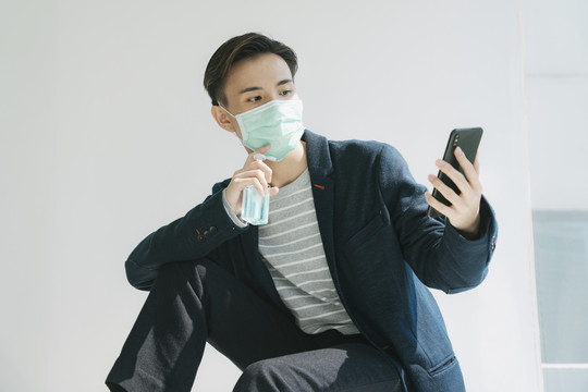 时尚写真-戴着医生面具的泰国亚裔年轻男子用智能手机和酒精凝胶清洗。