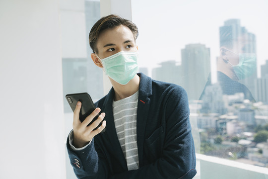 家中检疫概念——戴医生口罩的亚泰年轻男子用智能手机在hone做生意。