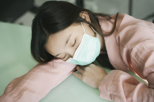 病人美丽的亚洲女人戴着医生的口罩，穿着粉色衬衫，躺在医院的桌子上等待医生。