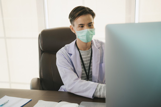 帅气的亚泰医生戴着口罩在工作台的房间里工作。在电脑上工作的医生正在研究治愈covid-19病毒的疫苗