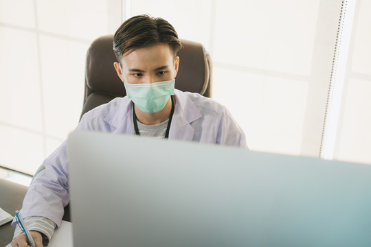 帅气的亚泰医生戴着口罩在工作台的房间里工作。在电脑上工作的医生正在研究治愈covid-19病毒的疫苗