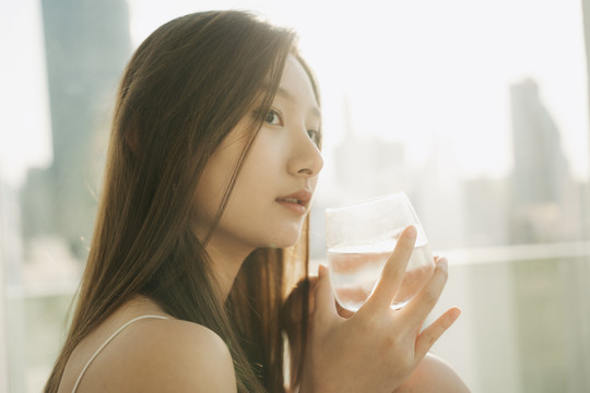 在摩天大楼的背景上，美丽的亚泰女人拿着一杯水的特写镜头。阳光。