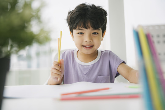 穿着紫色t恤的小男孩坐在靠窗的桌子旁，用铅笔和颜料在纸上画画和着色。