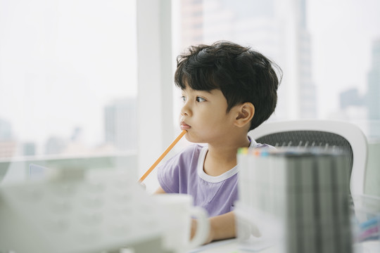 穿着紫色t恤的小男孩坐在靠窗的桌子旁，想着下一步要画什么。
