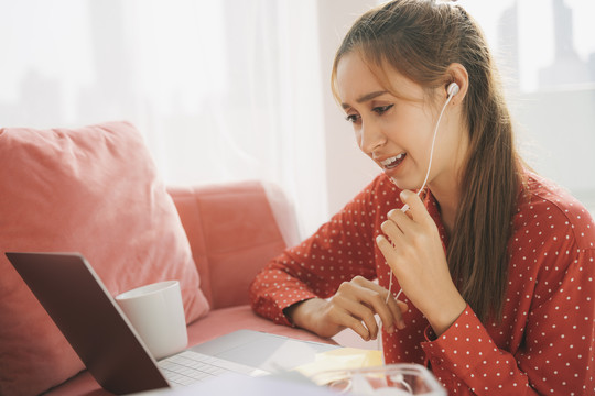 在家工作的概念-穿着红色睡衣衬衫的美丽年轻女子使用耳机享受与朋友在笔记本电脑上交谈。放松。检疫。