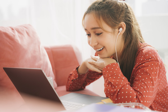 在家工作的概念-穿着红色睡衣衬衫的美丽年轻女子使用耳机享受与朋友在笔记本电脑上交谈。放松。检疫。