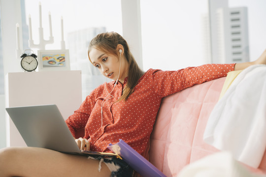 在家工作的概念-穿着红色睡衣衬衫的漂亮年轻女子坐在沙发上，用笔记本电脑的耳机听歌曲。在家工作。检疫。