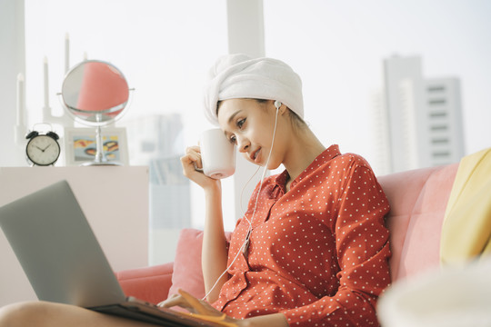 在家工作的概念-穿着红色睡衣衬衫，头上裹着毛巾的漂亮年轻女子坐在沙发上，拿着一杯咖啡，在笔记本电脑上