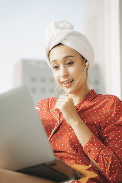 在家工作的概念-美丽的年轻女子穿着红色睡衣衬衫和毛巾浴巾头包，用笔记本电脑的耳机和朋友聊天。