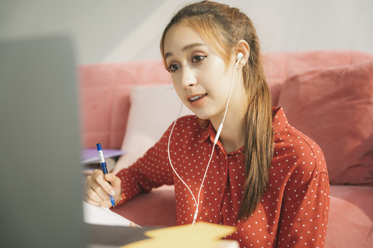 在家工作的概念-穿着红色睡衣衬衫、戴着耳机的漂亮年轻女子在家里用笔记本电脑与伴侣视频通话。检疫。