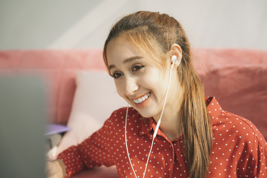 在家工作的概念-穿着红色睡衣衬衫的美丽年轻女子在笔记本电脑上与同事交谈。在家工作。使用耳机。检疫。