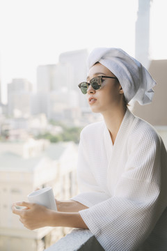 一位身穿浴衣、戴着墨镜的年轻女子在阳台上端着一杯白咖啡。