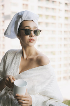 一位身穿白色浴袍、戴着墨镜的年轻女子在公寓房间的阳台上端着一杯咖啡。