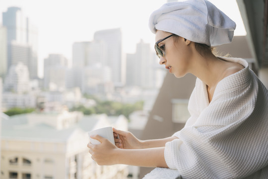 复制空间。侧视图-在公寓阳台，穿着白色浴袍的年轻女子拿着一杯咖啡。