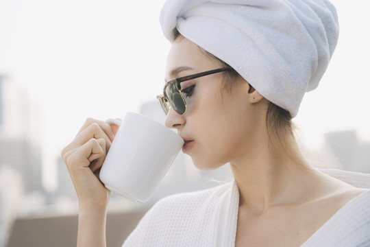 侧视图-年轻女子穿着白色浴袍，戴着墨镜，在家里的公寓里喝热咖啡。