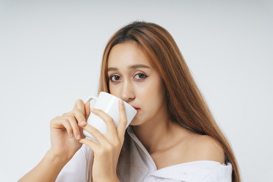 穿着白色衬衫的美丽女人，自然的妆容与白色背景隔离开来。护肤面部护理。喝白杯子里的水咖啡茶的女人。拿着