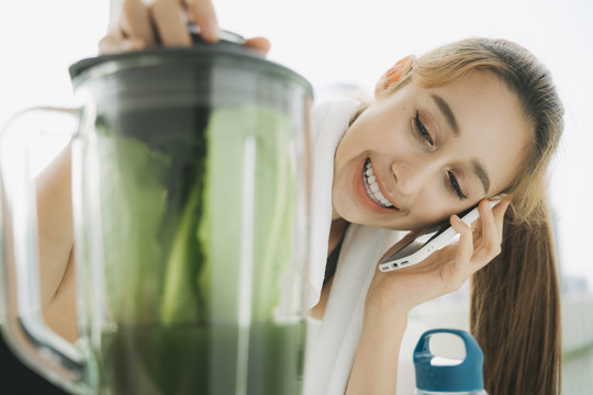 美丽健康的女人用搅拌机做健康蔬菜汁，在厨房里和朋友通电话。