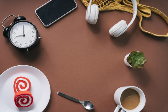固定的。平铺的甜点，热咖啡，勺子，时钟，桌子，耳机，智能手机在棕色的桌子上。