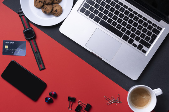 顶视图-办公桌工作场所，带有咖啡杯、饼干、智能手机、信用卡和笔记本电脑，背景为红色和黑色。