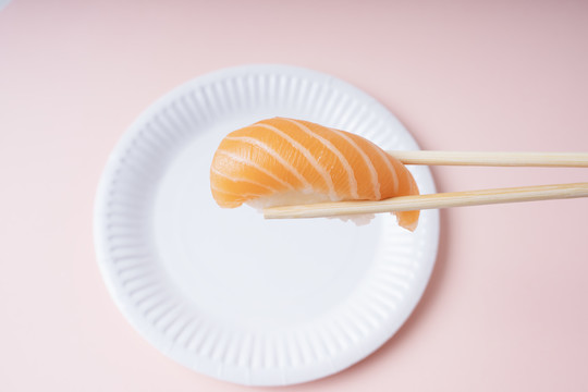 新鲜三文鱼寿司放在白色盘子里，用筷子夹在粉色背景上吃。