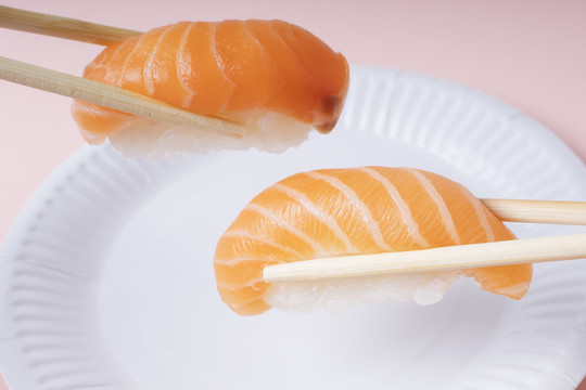 新鲜三文鱼寿司放在白色盘子里，用筷子夹在粉色背景上吃。
