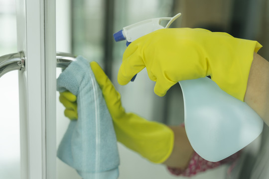 女佣戴着黄色防护手套，用抹布和洗涤剂喷雾瓶清洁门口。Covid-19冠状病毒预防。