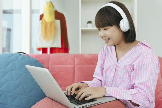 年轻的亚洲学生黑色短发女子戴着耳机在客厅的沙发上用笔记本电脑在家学习。