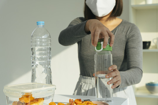 戴着白色口罩的女人在厨房里从瓶子里倒水。